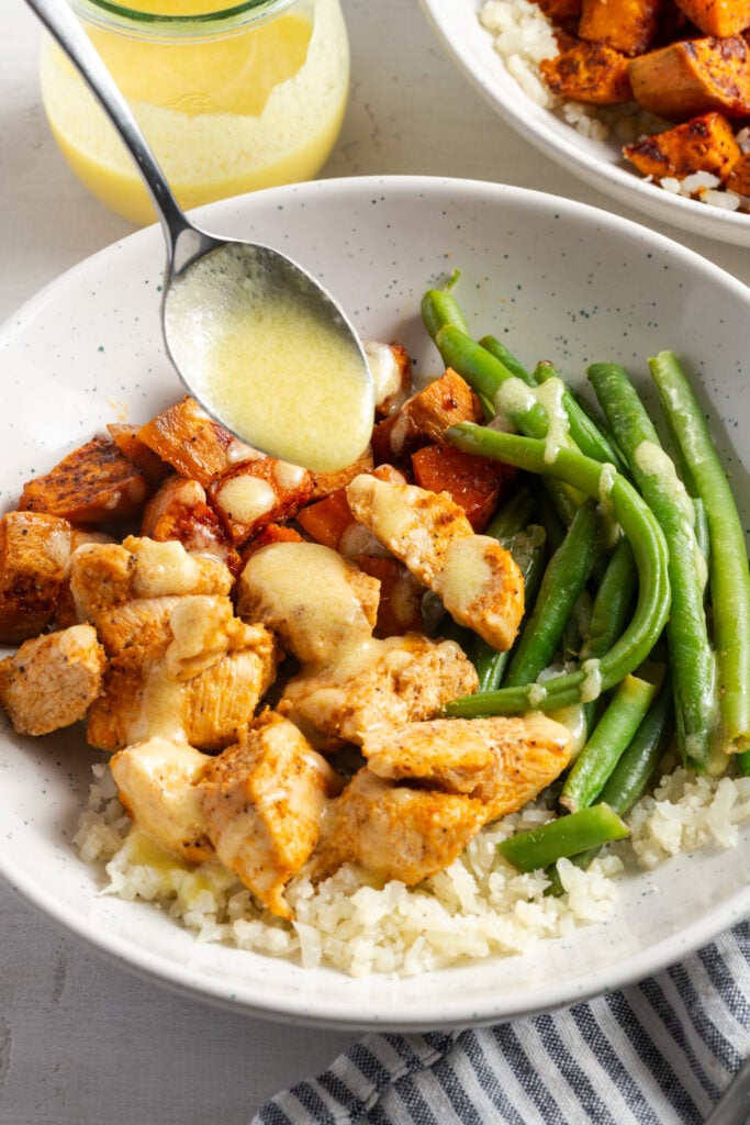 action shot of a spoon drizzling the garlic mojo sauce over a plated chicken sweet potato bowl.