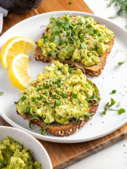 healthy avocado and egg salad on toasted multigrain on a white plate atop a wooden cutting board.
