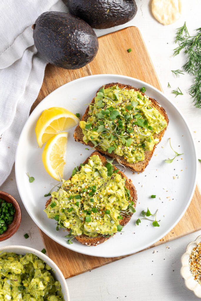 open faced avocado egg salad sandwiches on a white plate with lemon wedges.