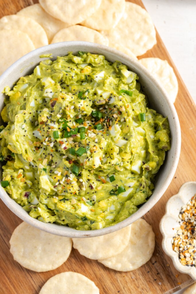 overhead shot of a white bowl of egg and avocado salad topped with everything but the bagel seasoning on a wooden cutting board with round rice crackers and a white pinch bowl of EBBB seasoning.