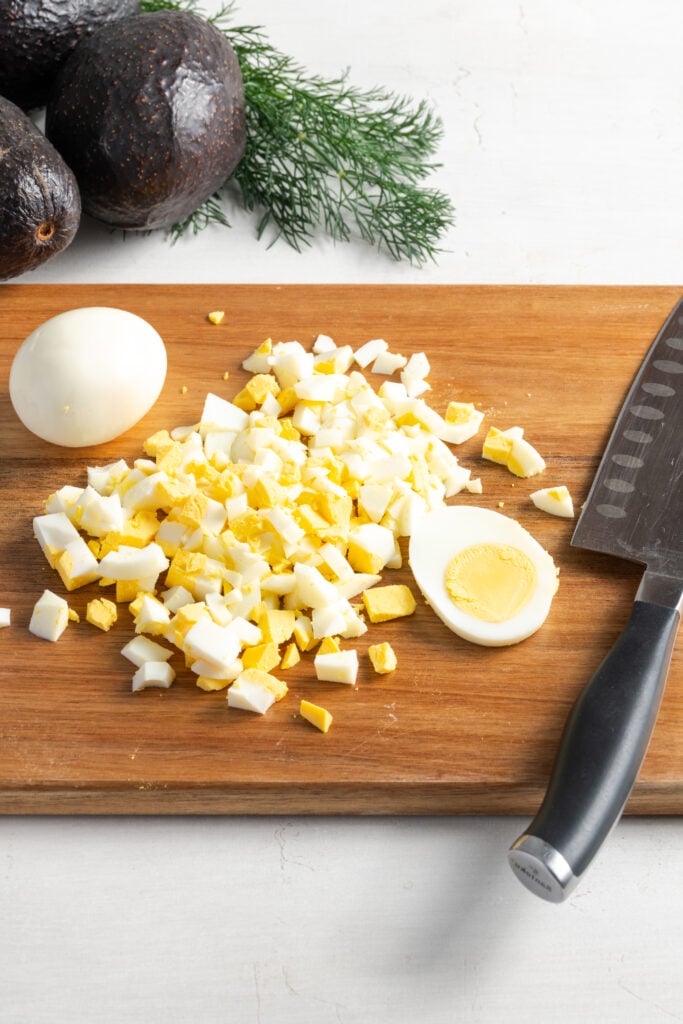 wooden cutting board with chopped boiled eggs on a white table with whole avocados and a sprig of fresh dill in the top left corner of the frame.