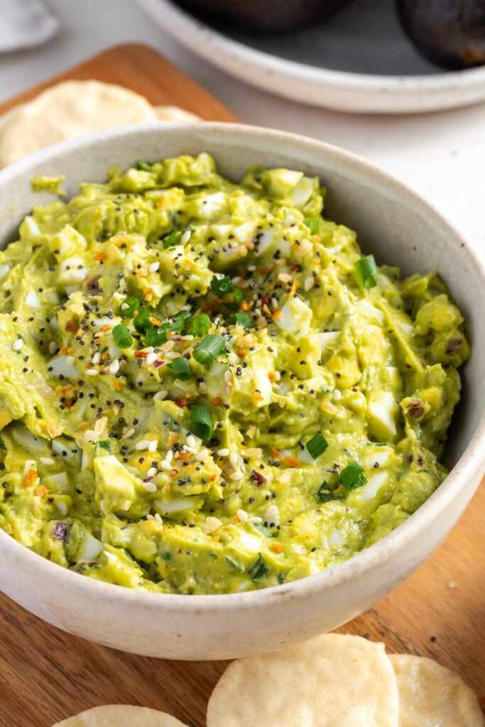 45 degree overhead shot of avocado egg salad topped with everything but the bagel seasoning on a wooden cutting board with round chips for dipping.