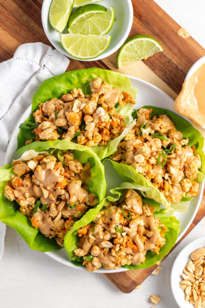 overhead shot of 4 healthy Asian ground turkey lettuce wraps on a white plate on a wooden cutting board with a white pinch bowl of lime wedges and a white pinch bowl of chopped peanuts.