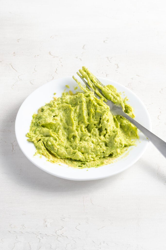 smashed avocado on a white plate with a silver fork.