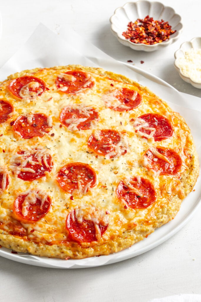 45 degree angle shot of a whole chicken crust pizza on a white plate with a bowl of red pepper flakes.