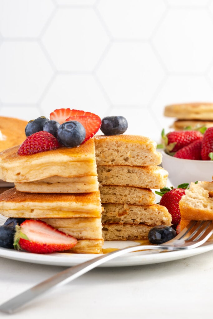 healthy cottage cheese pancakes topped with syrup and berries on a white plate with a bite taken out to reveal the fluffy interior.