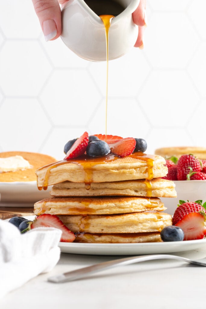 action shot of syrup being poured on a stack of cottage cheese pancakes with fresh berries on top.