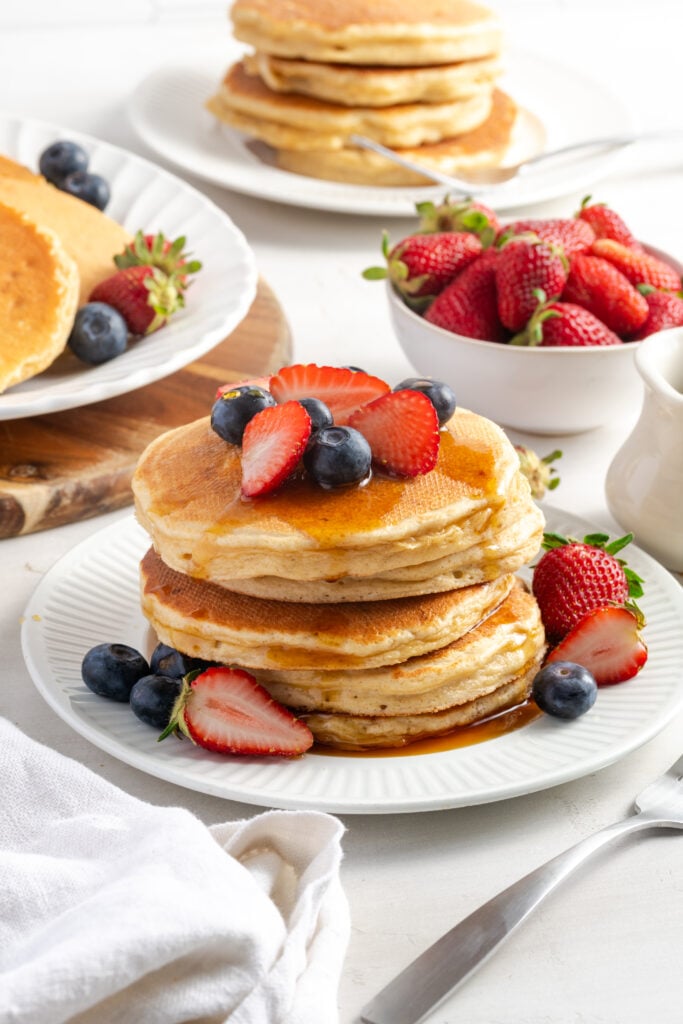 stack of high protein cottage cheese pancakes with syrup and berries with another plate with a stack of pancakes in the background without any toppings.