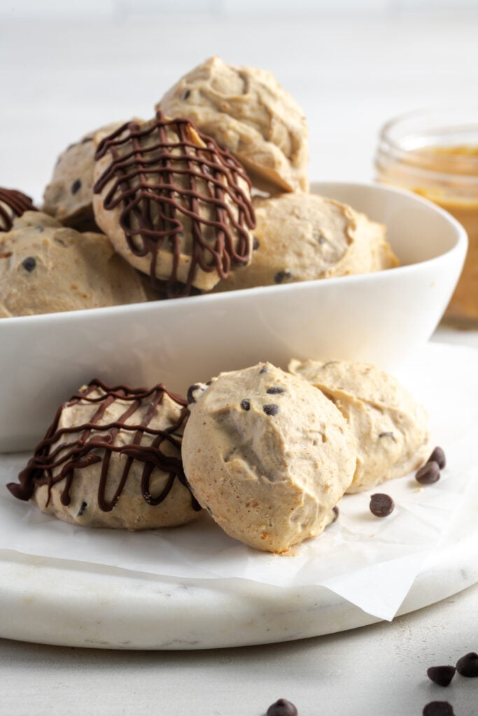 side view of Greek yogurt peanut butter balls in a white bowl and some on the table in front of the bowl with a few mini chocolate chips scattered around and a jar of peanut butter in the background.
