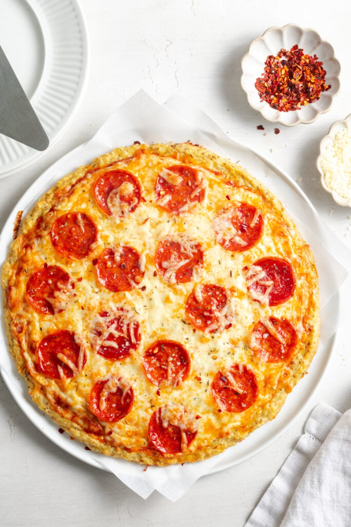overhead shot of pepperoni pizza with a chicken crust on a white plate with a pinch bowl of red pepper flakes and a pinch bowl of parmesan to the side.