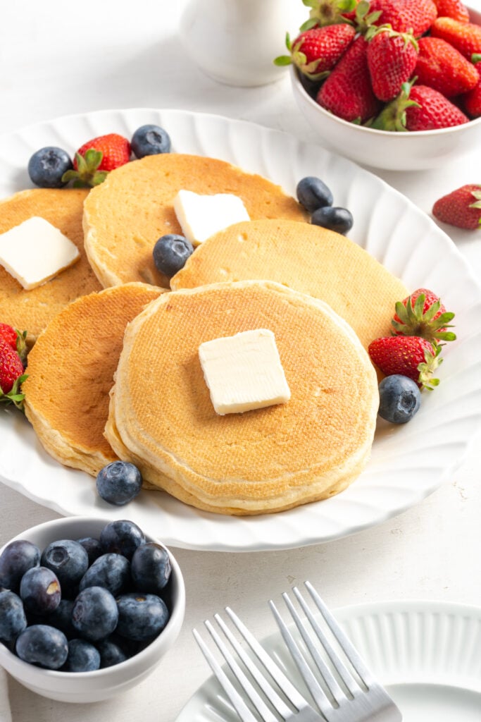 white plate of fluffy cottage cheese pancakes with pads of butter and fresh berries with a bowl of blueberries on one side and a bowl of strawberries on the other.