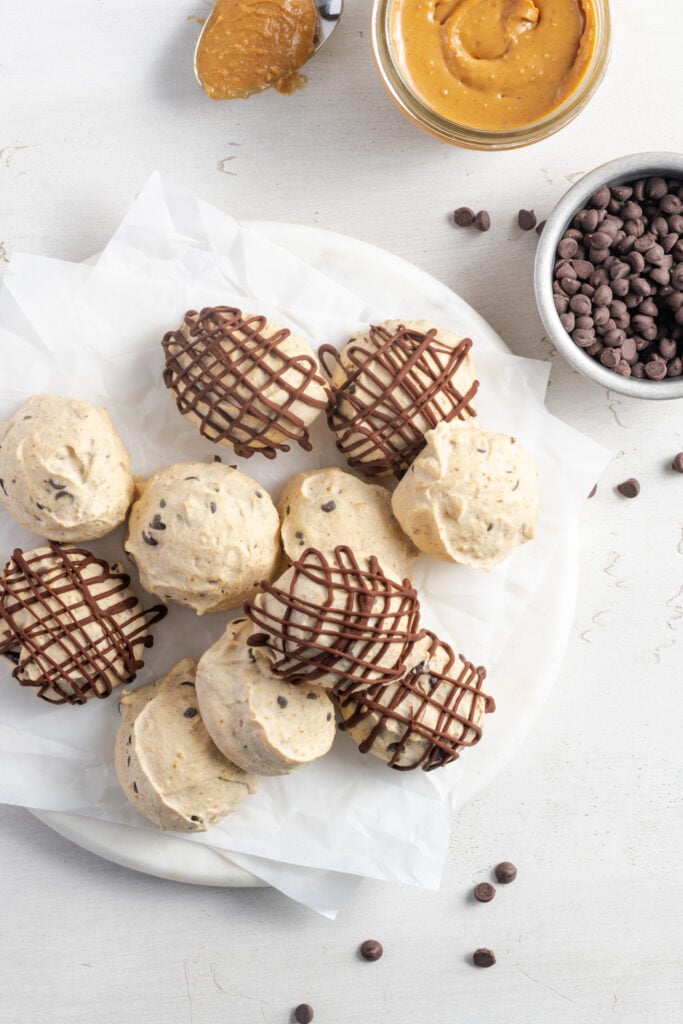 plate of creamy peanut butter frozen yogurt balls with half of them drizzled with melted chocolate on a white plate with a small bowl of mini chocolate chips and a small bowl of peanut butter.