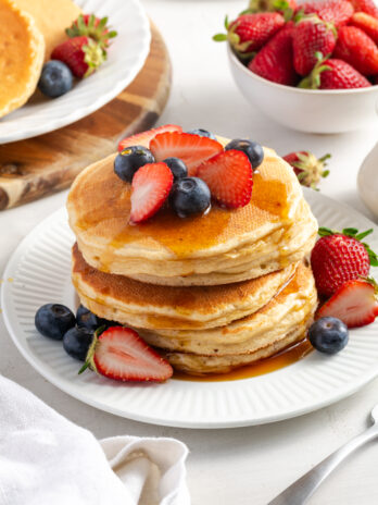 stack of cottage cheese pancakes with syrup and fresh berries on a white plate on a white table with a bowl of fresh strawberries and a plate of undressed pancakes.