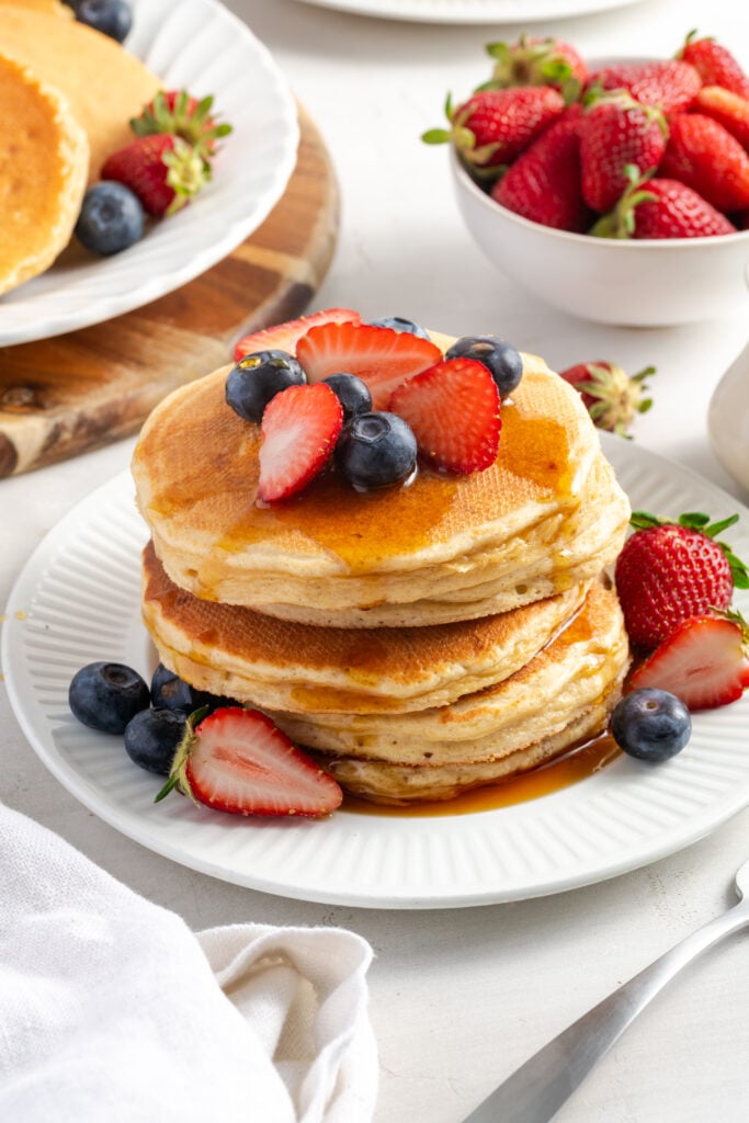 stack of cottage cheese pancakes with syrup and fresh berries on a white plate on a white table with a bowl of fresh strawberries and a plate of undressed pancakes.
