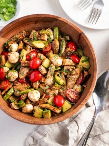 flat lay shot of a wooden bowl filled with chicken caprese salad on a white table with a silver serving spoon resting on top of a grey linen napkin, white dinner plates with silver forks, and a pinch bowl of extra basil leaves.
