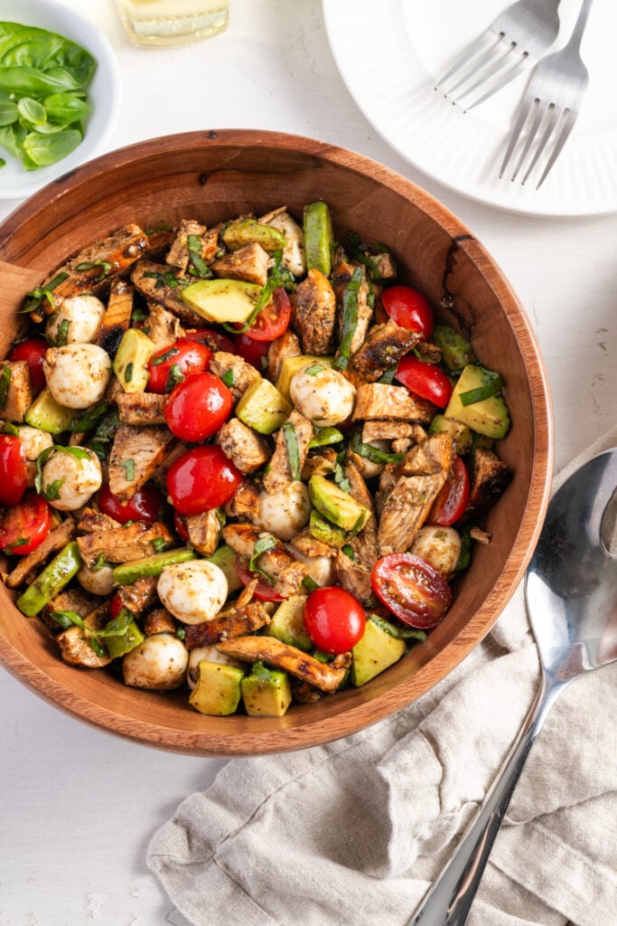 flat lay shot of a wooden bowl filled with chicken caprese salad on a white table with a silver serving spoon resting on top of a grey linen napkin, white dinner plates with silver forks, and a pinch bowl of extra basil leaves.