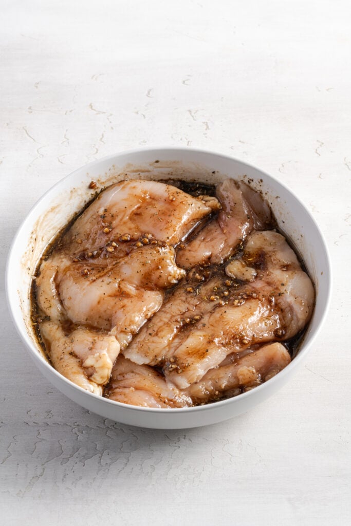 chicken breasts marinating in balsamic marinade in a white bowl on a white table.