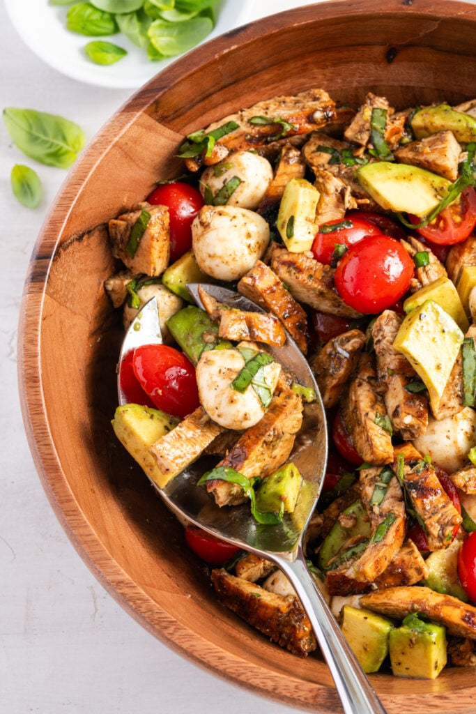 grilled chicken caprese salad in a wooden salad bowl with a large silver serving spoon taking a portion out.