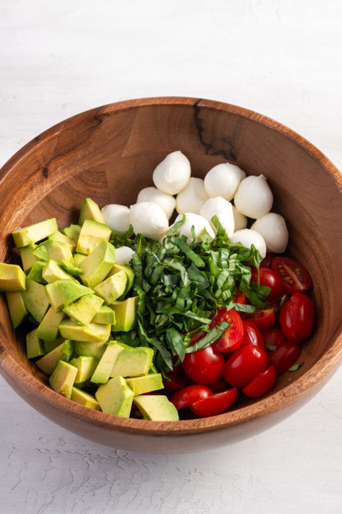 avocado, halved cherry tomatoes, fresh mozzarella balls, and chiffonade basil in a wooden mixing bowl.