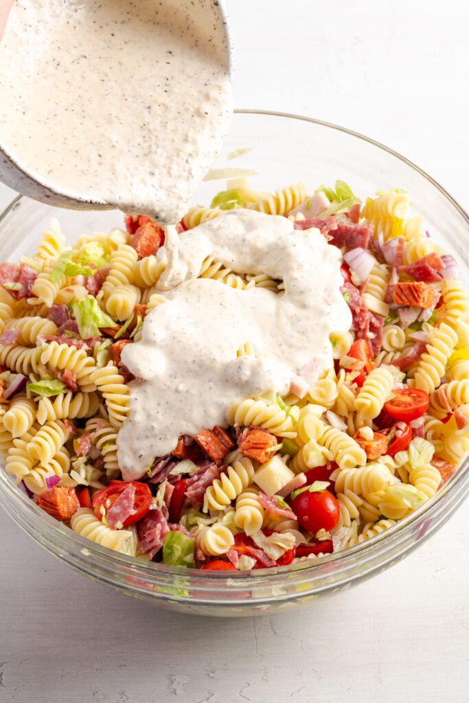 action shot of a hand pouring the creamy dressing over the bowl with the grinder pasta salad ingredients.