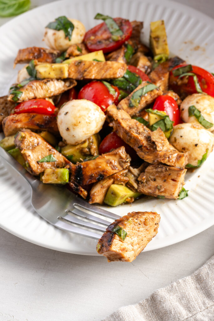 fork taking a bite of healthy grilled chicken caprese salad from a white dinner plate.