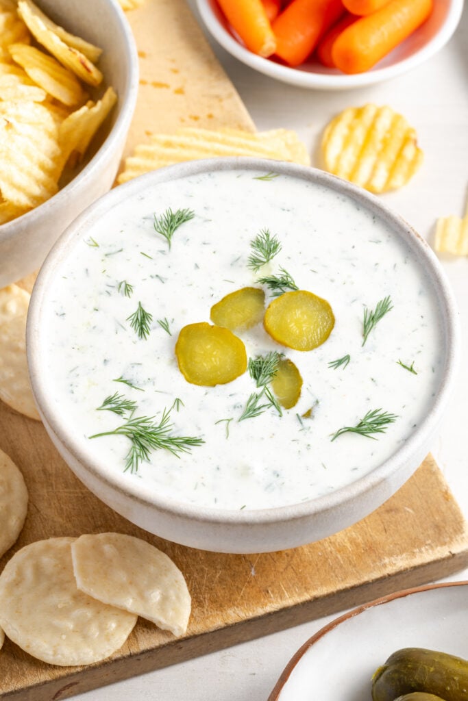 dill pickle cottage cheese and greek yogurt dip in a white bowl on a wooden snack board; the top of the dip is garnished with fresh dill leaves and sliced pickles.