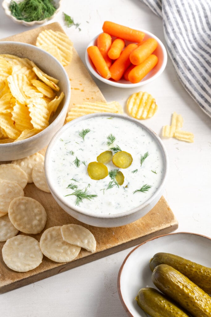 snack board with a bowl of high protein dill pickle dip garnished with fresh dill and pickle slices, a bowl of ridged potato chips, rice crackers, baby carrots, and a side of pickles.