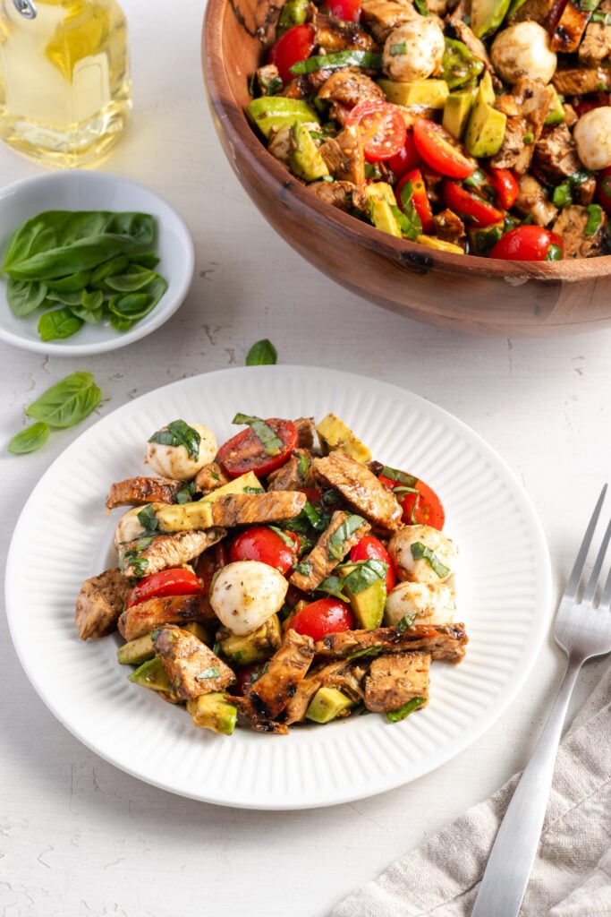 white plate of caprese chicken salad on a. white table with a silver fork and grey linen napkin with a wooden salad bowl with the rest of the recipe and a pinch bowl of basil leaves framing the top of the shot.