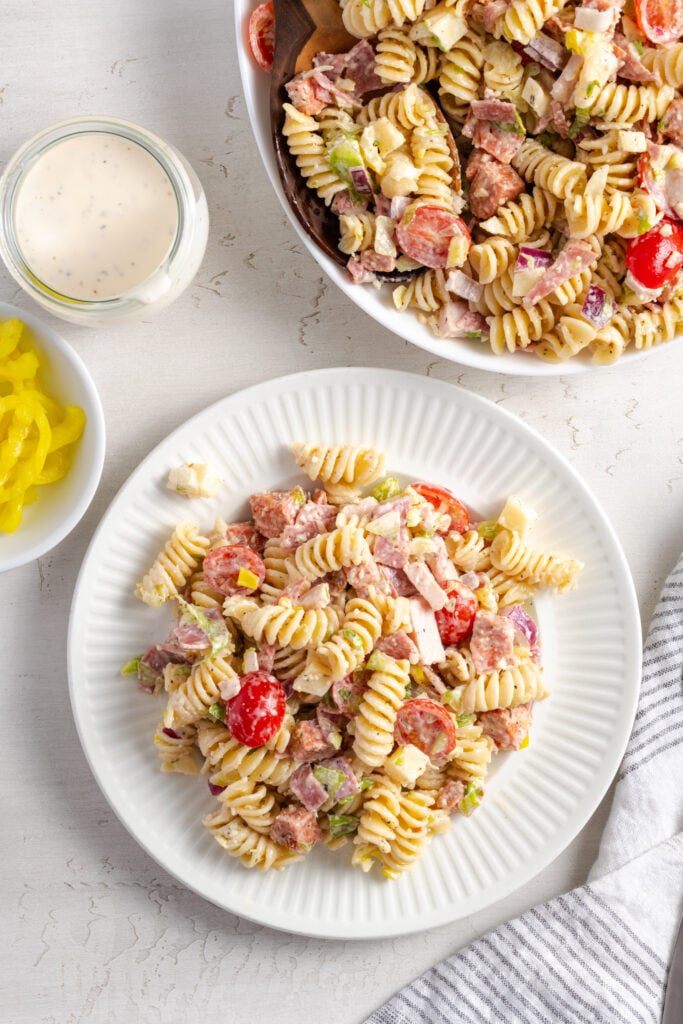 flat lay shot of a portion of Italian grinder pasta salad on a white dinner plate on a white table with a jar of extra dressing and a bowl of extra peppers to the side.