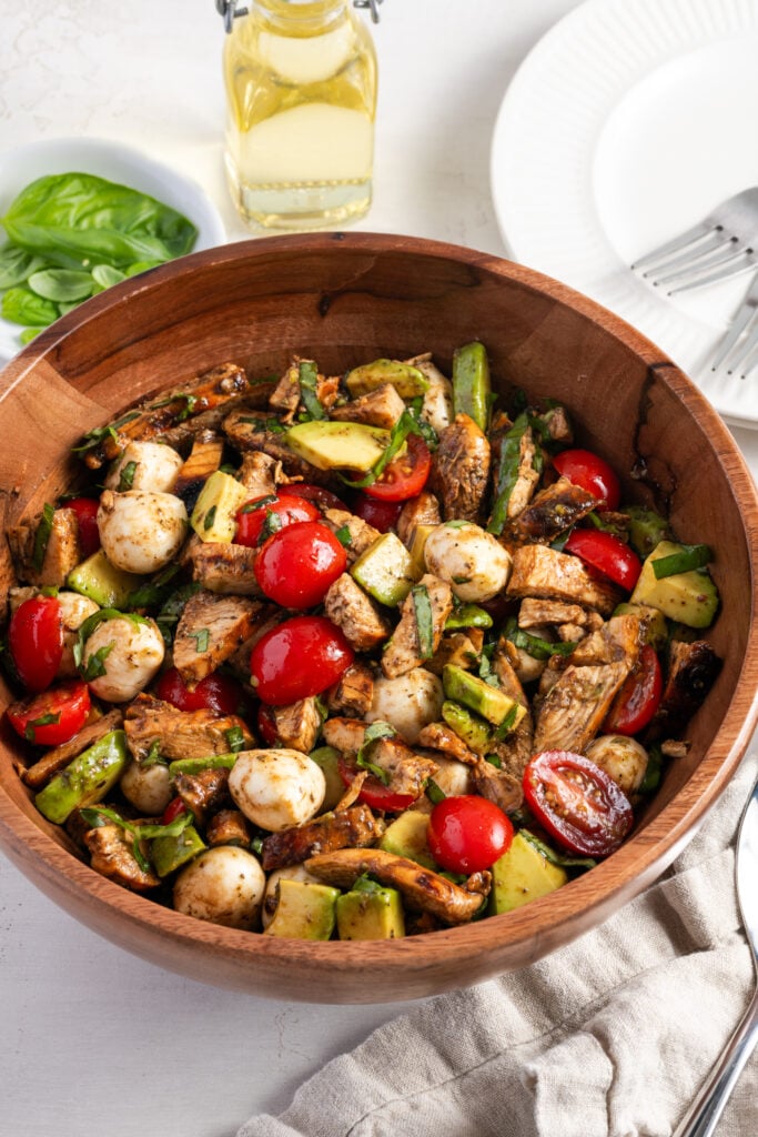 wooden bowl filled with chicken caprese salad with fresh avocado on a white table with a bowl of fresh basil leaves and a small jug of olive oil.