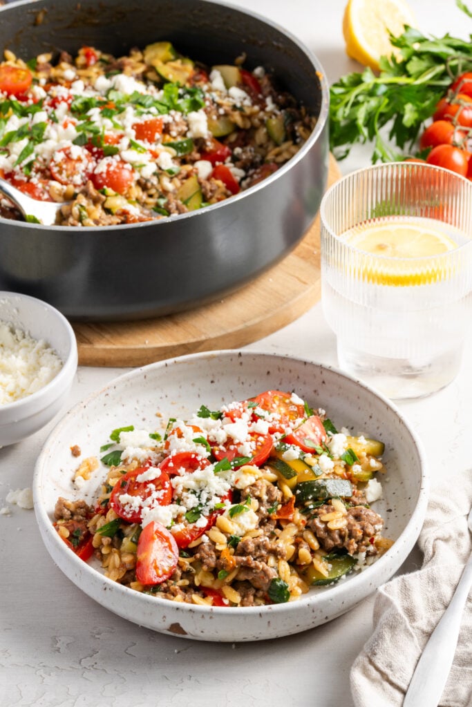 plated portion of greek beef and orzo skillet in a white earthenware pasta bowl with a glass of lemon water, the remaining skillet, and fresh produce in the background.