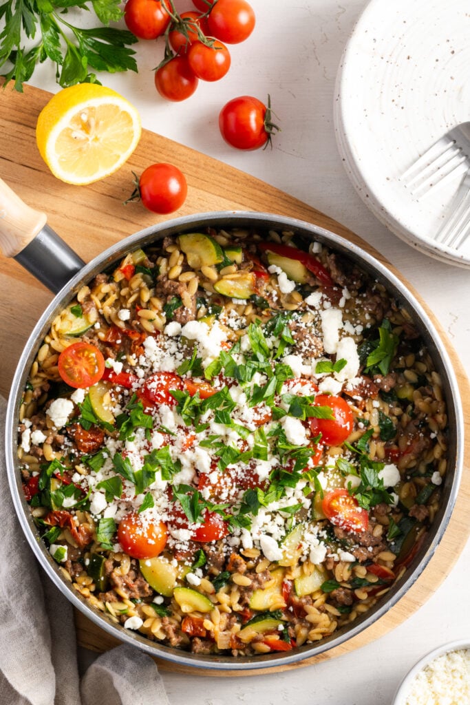 flat lay shot of a garnished greek ground beef and orzo skillet on a wooden trivet on a white table with fresh produce and white earthenware plates.