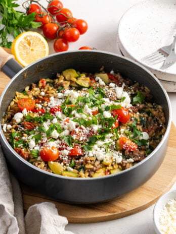 greek ground beef orzo skillet topped with fresh feta, herbs, and cherry tomatoes on a wooden trivet on a white table with a bunch of cherry tomatoes, a bunch of parsley and a halved lemon in the background.