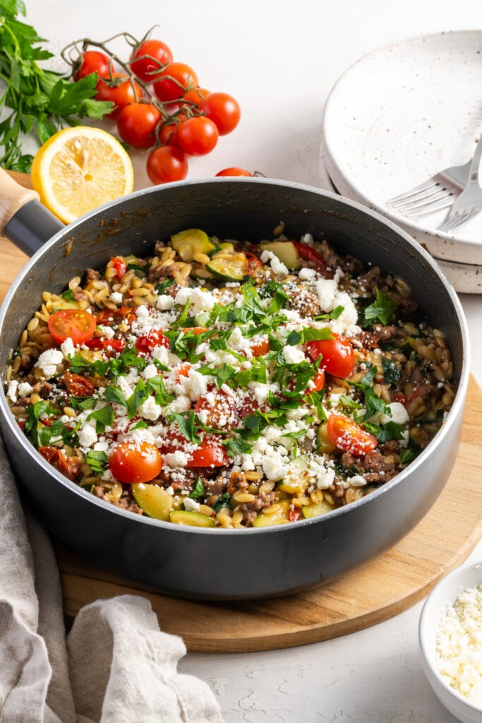 greek ground beef orzo skillet topped with fresh feta, herbs, and cherry tomatoes on a wooden trivet on a white table with a bunch of cherry tomatoes, a bunch of parsley and a halved lemon in the background.