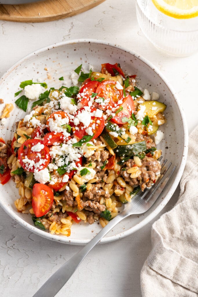 flat lay shot of a white earthenware pasta bowl filled with a portion of Greek ground beef orzo skillet on a white table with a glass of lemon water.