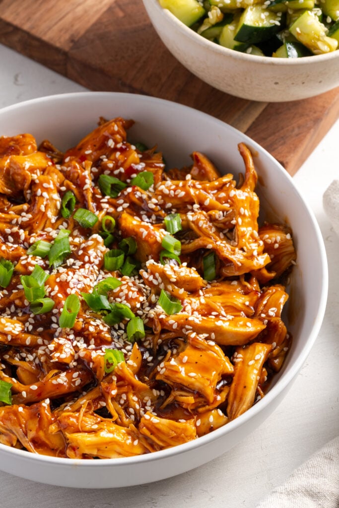 closeup shot of a bowl of saucy shredded sesame chicken topped with scallions and sesame seeds.