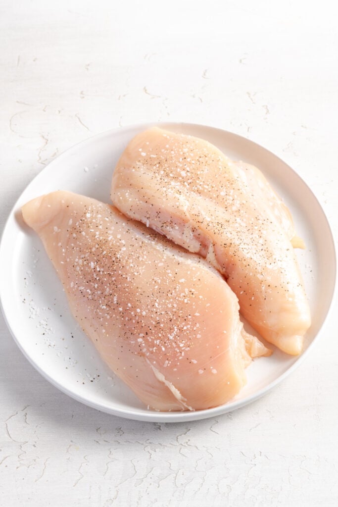 two raw, seasoned chicken breasts on a white plate.