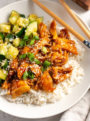 flat lay shot of a sesame chicken and rice bowl on a white table with a pair of wooden chopsticks.