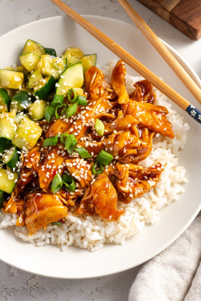 flat lay shot of a sesame chicken and rice bowl on a white table with a pair of wooden chopsticks.