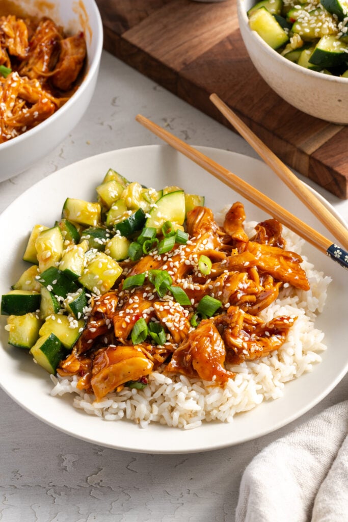 45 degree angle of a white pasta bowl filled with steamed rice, shredded sesame chicken breast, and cucumber salad with a pair of wooden chopsticks balancing on the side of the plate.