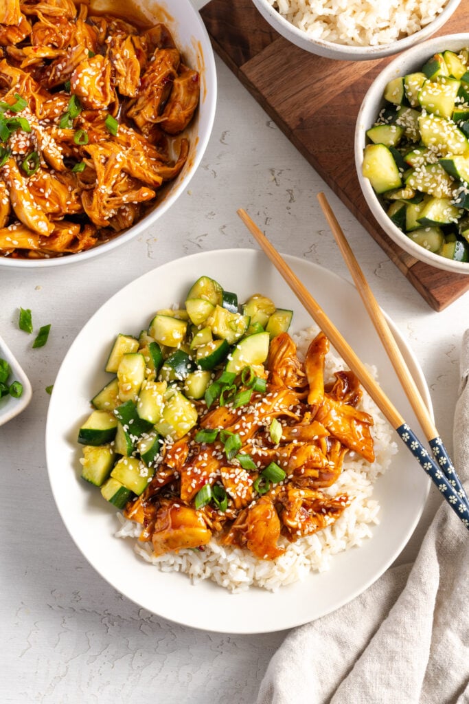 plated portion of shredded sesame chicken rice bowl on a white table with bowls of all the individual components — rice, chicken, cucumber salad, scallions —scattered around it.
