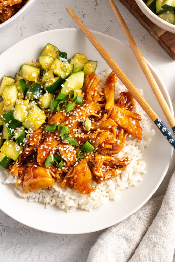 overhead closeup shot of a plate with a serving of shredded sesame chicken and rice with a cucumber salad and green onions as garnish.